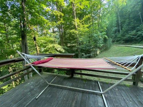 A colorful hammock is set up on a wooden deck, surrounded by lush green trees and a peaceful pathway.