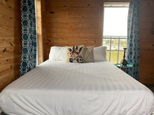 Cozy bedroom with a white bed, decorative pillows, and blue patterned curtains, featuring wooden walls and a side table.