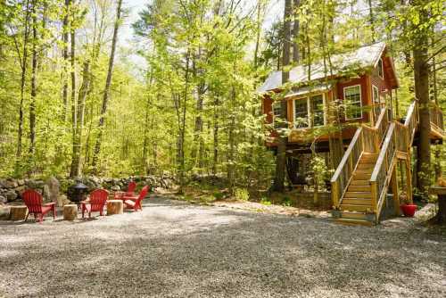 A cozy red cabin on stilts surrounded by lush green trees, with a gravel area and red chairs for outdoor seating.