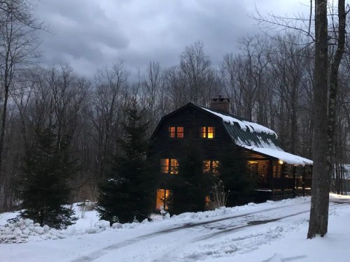 A cozy cabin in the snow, with warm lights glowing from the windows, surrounded by trees under a cloudy sky.