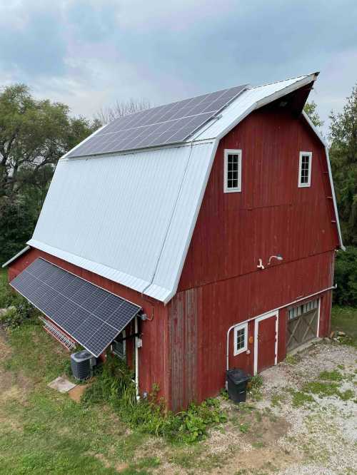 A red barn with a metal roof, featuring solar panels on top and plants growing around its base.