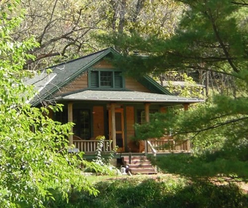 A charming wooden house surrounded by trees, featuring a porch and green shutters, nestled in a natural setting.