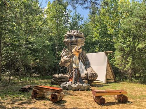 A large wooden sculpture of a creature holding a bird, surrounded by benches and a tent in a forested area.