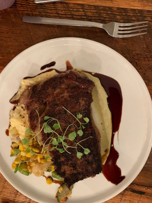 A plate of steak on creamy mashed potatoes, topped with microgreens and accompanied by corn and vegetables.