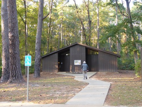 A person walks towards a wooden building surrounded by tall trees, with a wheelchair-accessible sign nearby.