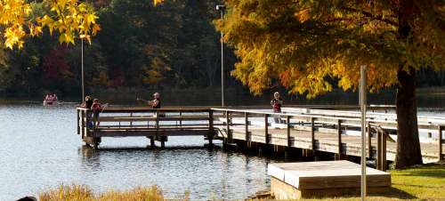 A wooden dock extends over a calm lake, with people fishing and enjoying the autumn foliage in the background.