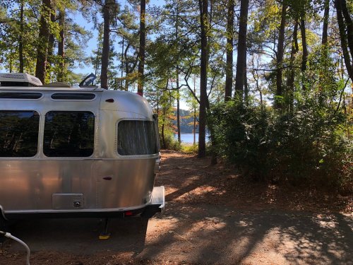 A shiny Airstream trailer parked near a wooded area, with a view of a lake in the background.