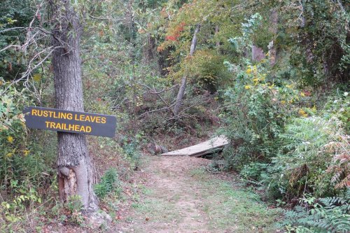 A trailhead sign reading "Rustling Leaves" next to a path surrounded by trees and greenery.