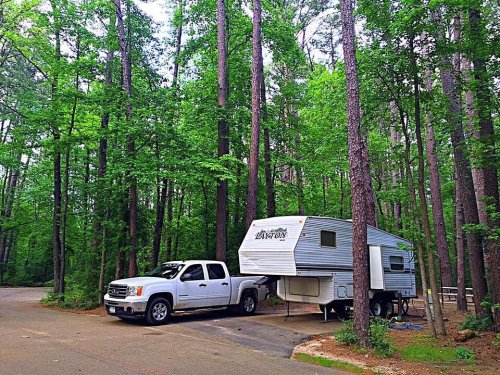 A white pickup truck parked next to a travel trailer in a lush, green forest setting.