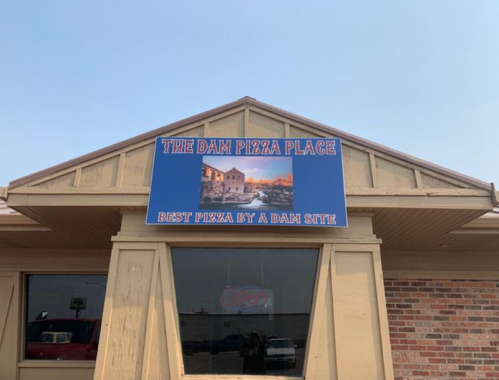 Sign for "The Dam Pizza Place" featuring the tagline "Best Pizza by a Dam Site" on a building exterior.