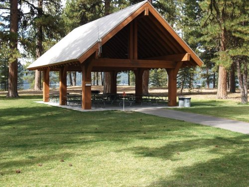 A wooden pavilion with a sloped roof, surrounded by trees and grass, featuring picnic tables underneath.