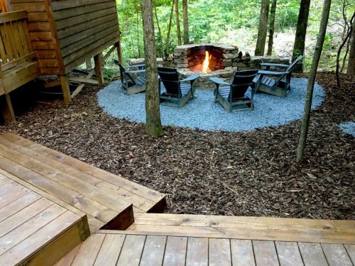 A cozy fire pit surrounded by gravel and wooden chairs, nestled in a wooded area with trees and a cabin nearby.