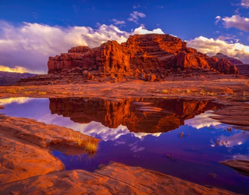 A vibrant sunset over a red rock formation, reflecting in a calm pool of water with clouds in the sky.