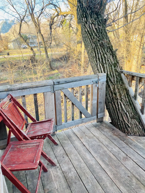 Two red wooden chairs on a rustic wooden deck, with a large tree trunk nearby and a peaceful natural setting.