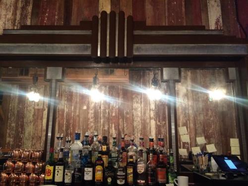 A rustic bar with a variety of liquor bottles displayed on shelves, illuminated by hanging lights against a weathered wall.