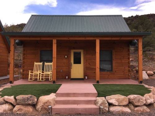 A cozy wooden cabin with a green metal roof, yellow door, and rocking chairs on the porch, surrounded by rocks and grass.