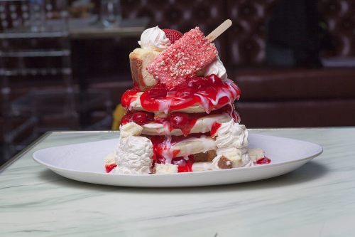 A towering dessert with layers of cake, whipped cream, strawberries, and a popsicle, drizzled with red sauce.