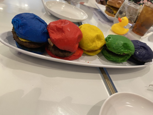 A platter of colorful burger sliders in red, blue, yellow, green, and purple buns, with a rubber duck and drinks in the background.