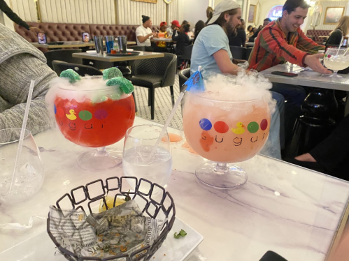 Two colorful, foggy cocktails in large bowls sit on a table, with a partially eaten basket of food nearby.