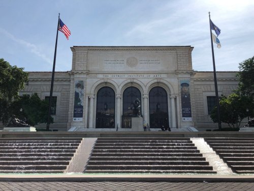 The Detroit Institute of Arts building with flags, steps, and a fountain in front under a clear blue sky.