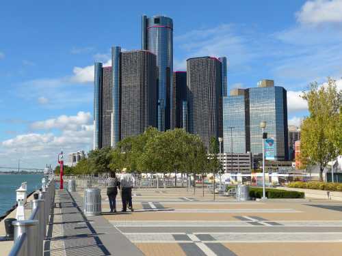 Two people walk along a waterfront promenade with modern buildings and a clear blue sky in the background.