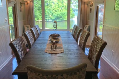 A spacious dining table with woven chairs, set near large windows overlooking a serene lake and greenery.