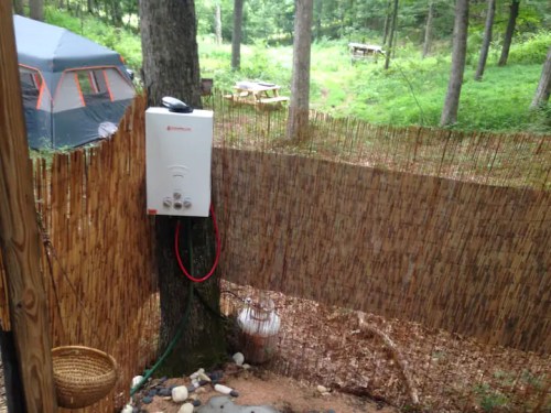 A portable water heater mounted on a tree, surrounded by a bamboo fence in a wooded camping area.