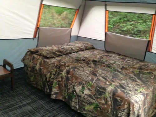 Interior of a tent featuring a camo-patterned bedspread and two pillows, with large windows letting in natural light.