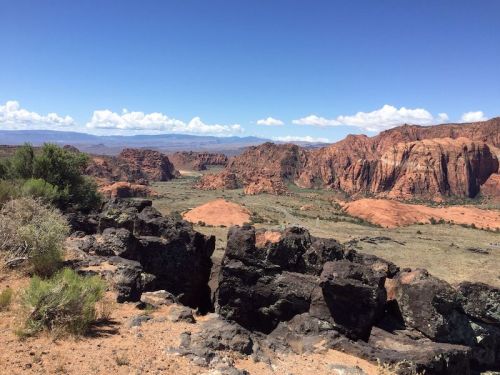A panoramic view of red rock formations and a vast landscape under a clear blue sky with scattered clouds.