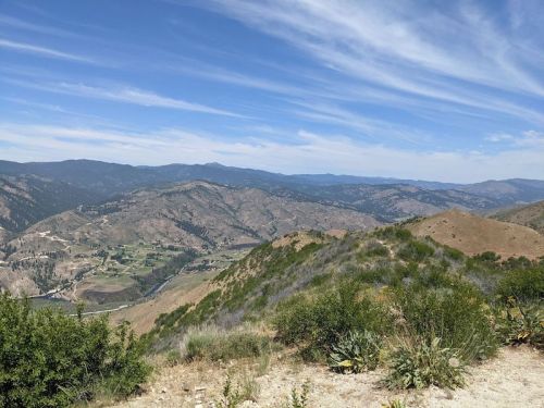 A panoramic view of rolling hills and valleys under a blue sky with wispy clouds. Green vegetation in the foreground.