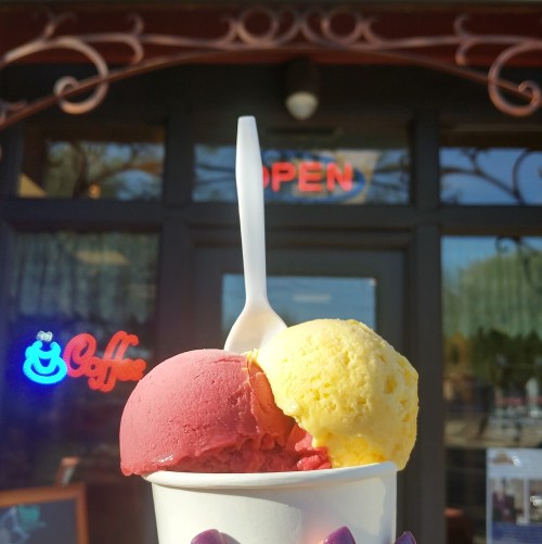 A cup of red and yellow ice cream with a spoon, in front of a shop with an "OPEN" sign.
