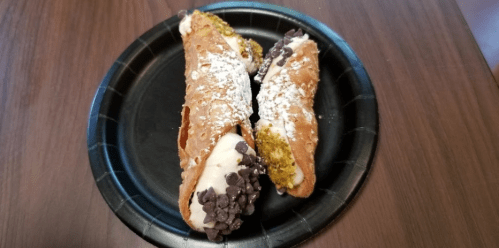 Two cannoli filled with cream, topped with chocolate chips and powdered sugar, served on a black paper plate.