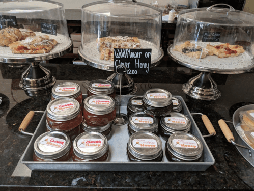 A display of jars of honey on a tray, with pastries in glass domes behind them. Prices and labels are visible.