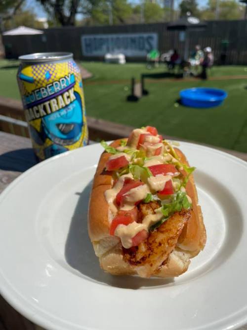 A hot dog topped with grilled chicken, tomatoes, and green onions, served on a plate with a can of blueberry soda.