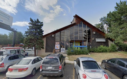 A modern building with large windows, surrounded by trees and parked cars, under a partly cloudy sky.