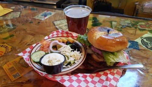 A burger with lettuce, tomato, and onion, served with a side salad and a pint of beer on a wooden table.