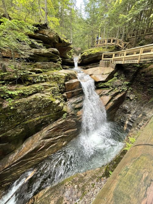A serene waterfall cascades over rocky cliffs, surrounded by lush greenery and wooden walkways.