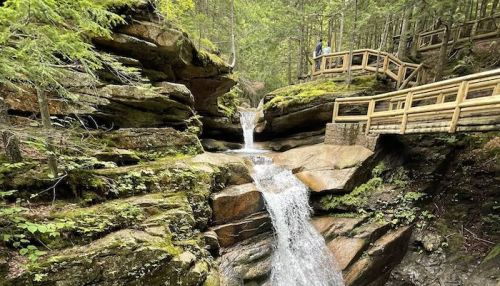 A serene waterfall cascades over rocks in a lush forest, with a wooden walkway and a person standing nearby.