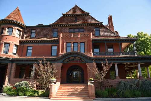 A large, ornate red brick mansion with a steep roof and decorative details, surrounded by greenery and blue sky.