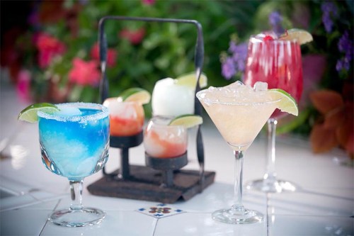 A colorful display of cocktails, including a blue drink, a margarita, and a red drink, with lime garnishes.