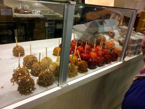 A display of caramel apples, some coated in nuts, at a candy shop. Various colors and toppings are visible.