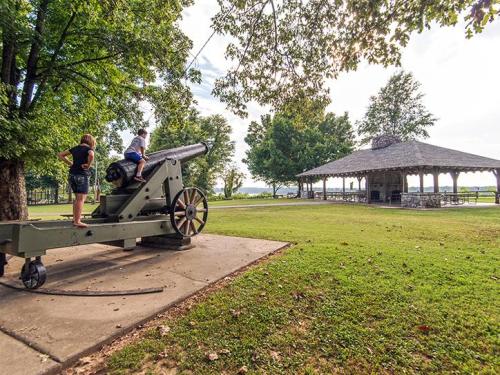 Two people explore a historic cannon near a gazebo, surrounded by trees and open green space.