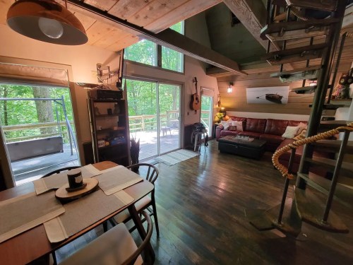 Cozy interior of a cabin with a dining area, living room, and spiral staircase, surrounded by greenery outside.