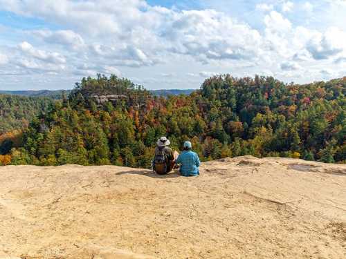 Two hikers sit on a rocky ledge, overlooking a colorful forest landscape under a partly cloudy sky.