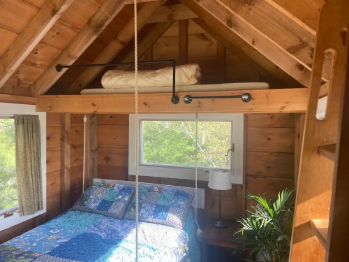 Cozy wooden bedroom with a lofted bed, colorful quilt, and a small lamp beside a window. Greenery visible outside.
