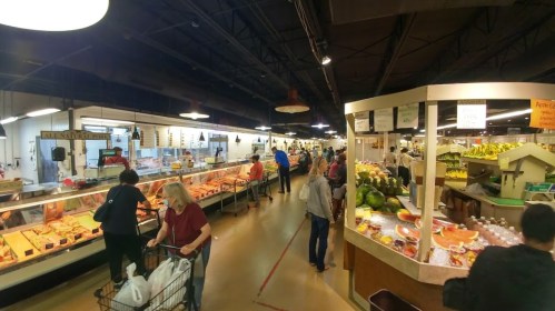 A bustling market scene with shoppers browsing various food stalls, including fresh produce and meats.