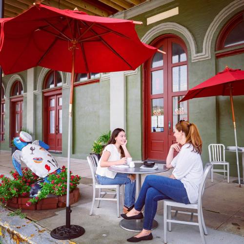 Two women sit at a table under red umbrellas, enjoying coffee outside a charming café with colorful decor.
