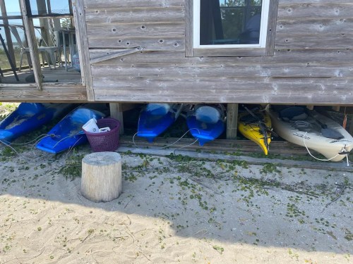 A wooden structure with kayaks stored underneath, including blue and yellow kayaks, on a sandy area.