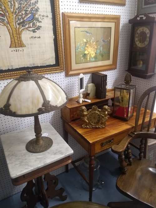 A cozy vintage display featuring a lamp, wooden desk, clock, framed art, and various antique decor items.