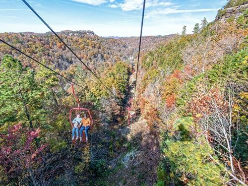 Two people ride a chairlift over a colorful autumn landscape with trees and hills in the background.